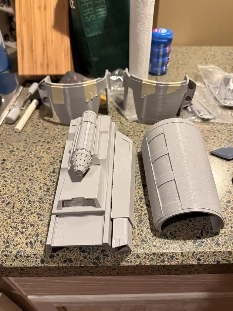 Several gray 3D-printed parts, including a detailed lightsaber hilt and curved pieces, are laid out on a speckled kitchen…