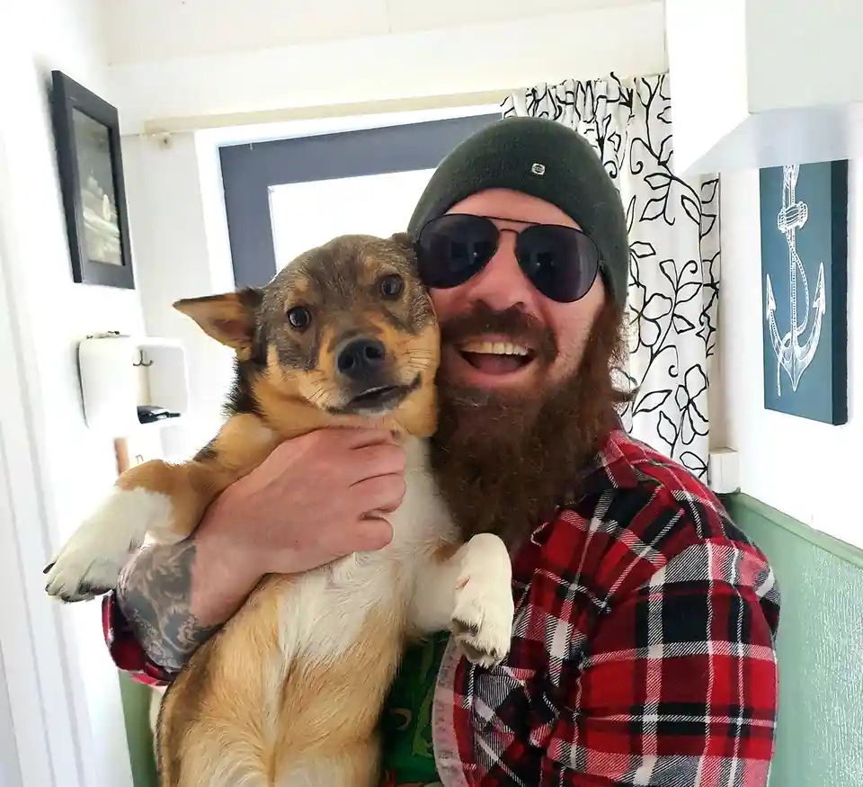 A man with a long beard, sunglasses, and a green beanie is smiling broadly while holding a tan and white dog. The dog looks…