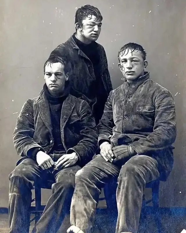 A vintage black-and-white photograph shows three young men in worn, dirty work clothes posing for a portrait. One stands…