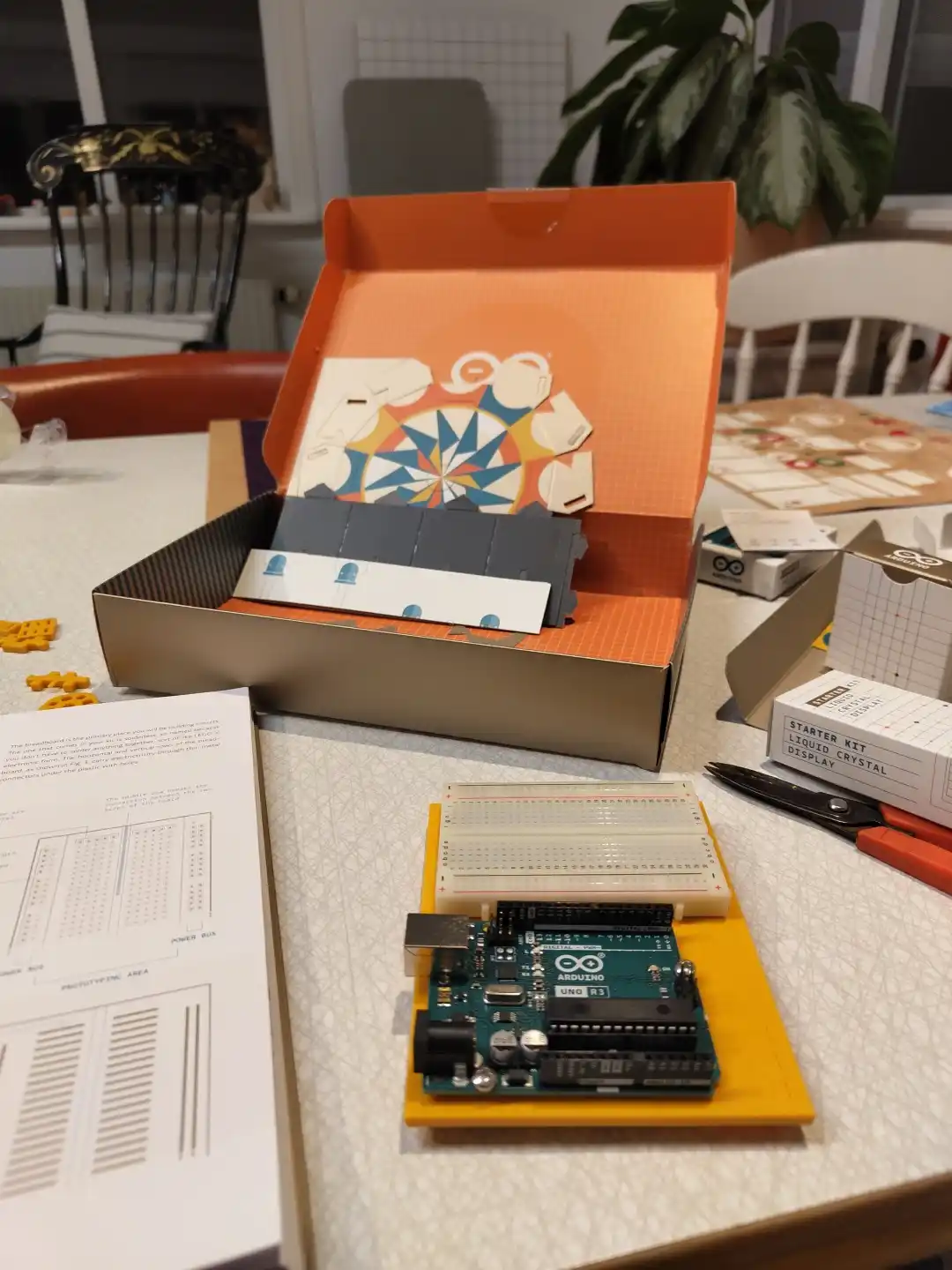 An open orange Arduino starter kit box sits on a table, revealing its contents including a breadboard, jumper wires, and an…
