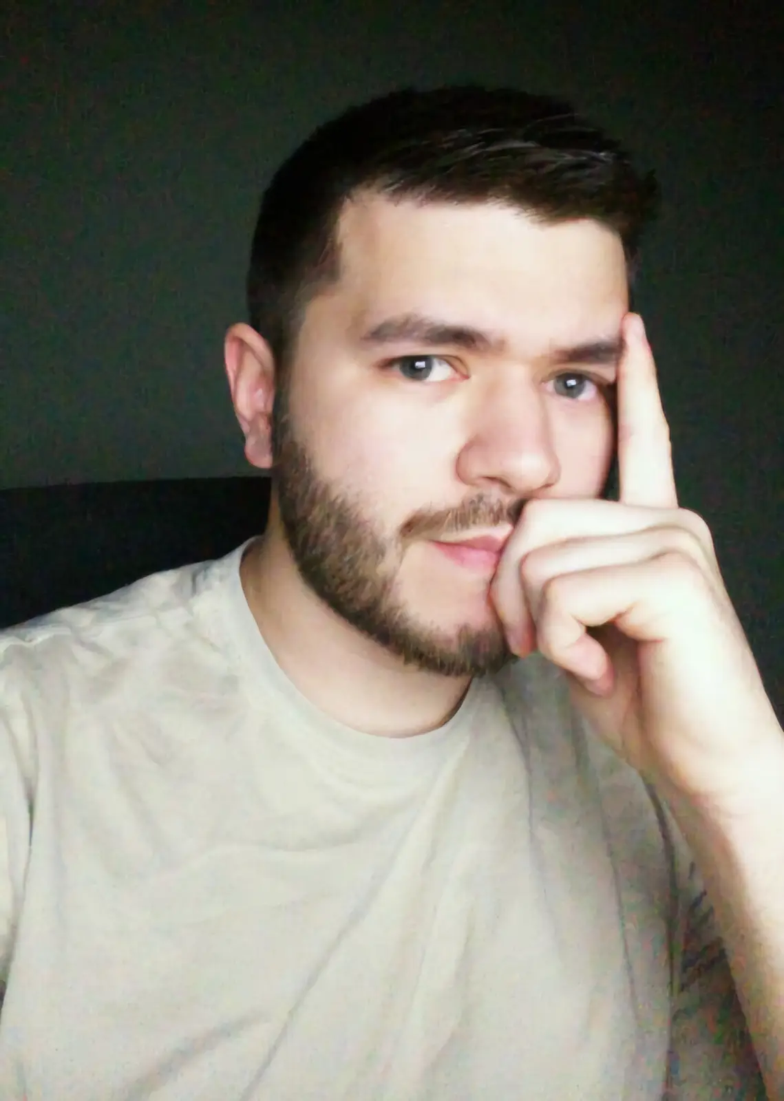 A man with a beard and short dark hair, wearing a light-colored t-shirt, looks directly at the camera with a slightly smug…