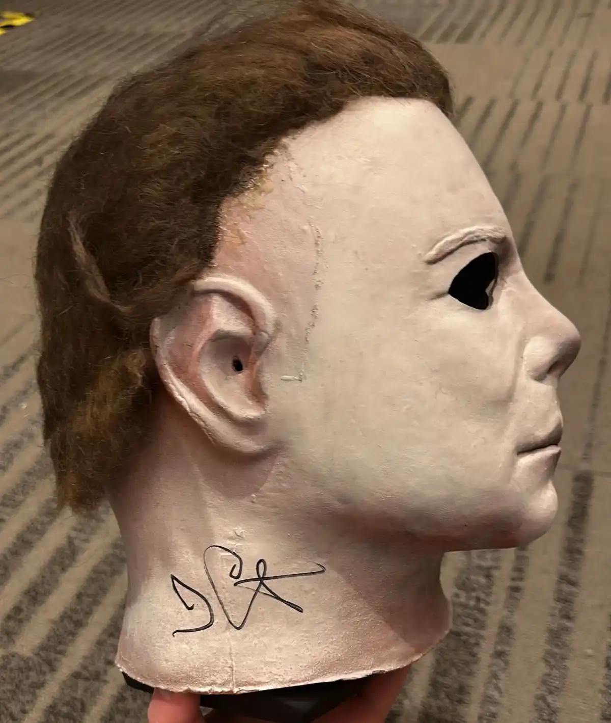 A close-up photo shows a white, featureless Michael Myers mask from the Halloween franchise, viewed from the side. The mask…