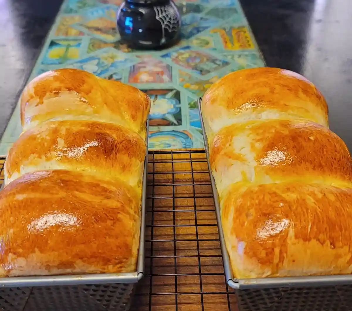 Two golden-brown, glossy loaves of bread sit side-by-side in metal loaf pans on a cooling rack, placed on a colorful…