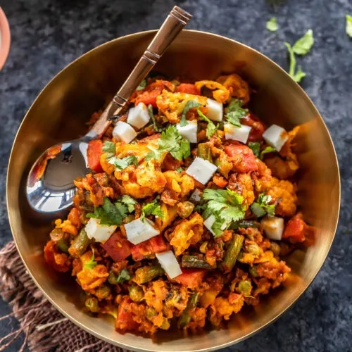 a top-down photo of a golden bowl filled with a colorful Indian-style curry dish, garnished with chopped cilantro and white…