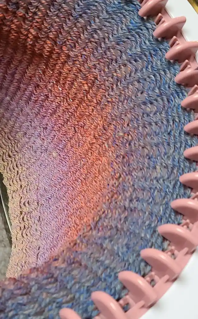 A close-up photo shows a colorful, gradient-knit fabric on a pink circular knitting loom, transitioning from warm…