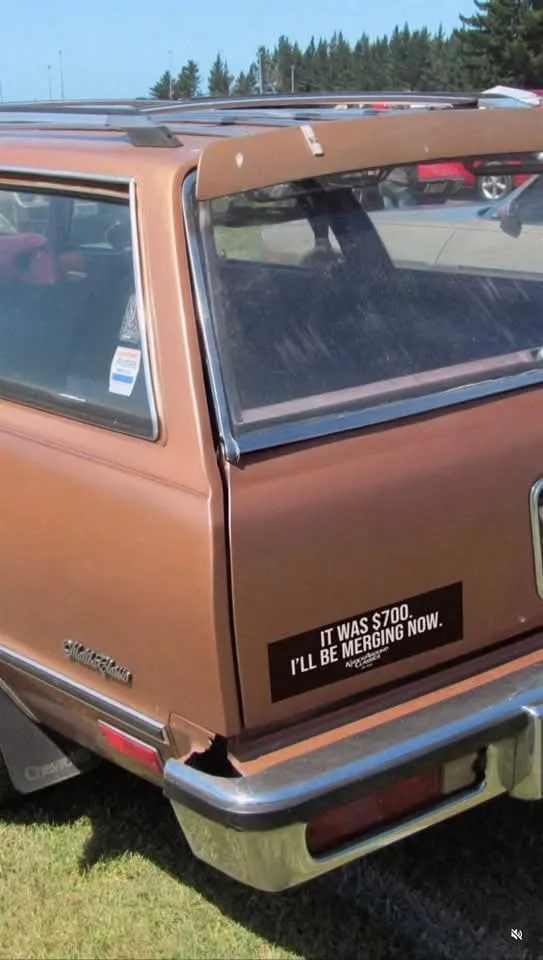 A rear view of a rusty brown 1970s Chevrolet Suburban station wagon parked outdoors. A black sticker on the rear quarter…