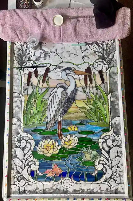An overhead photo of a partially completed stained glass art project depicting a heron standing in water with lily pads and…