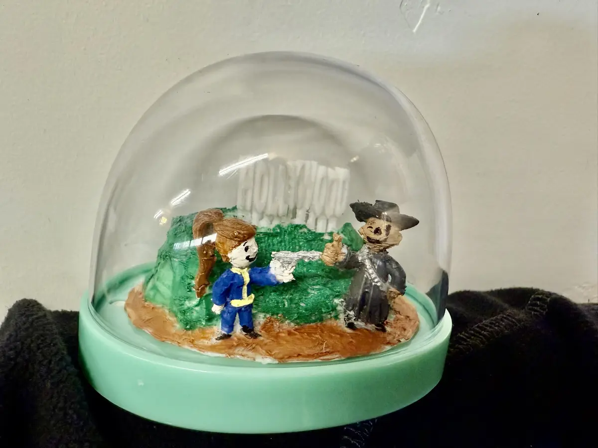 A handmade diorama under a glass dome shows two clay figures: one in a blue jumpsuit holding a silver gun, facing another…
