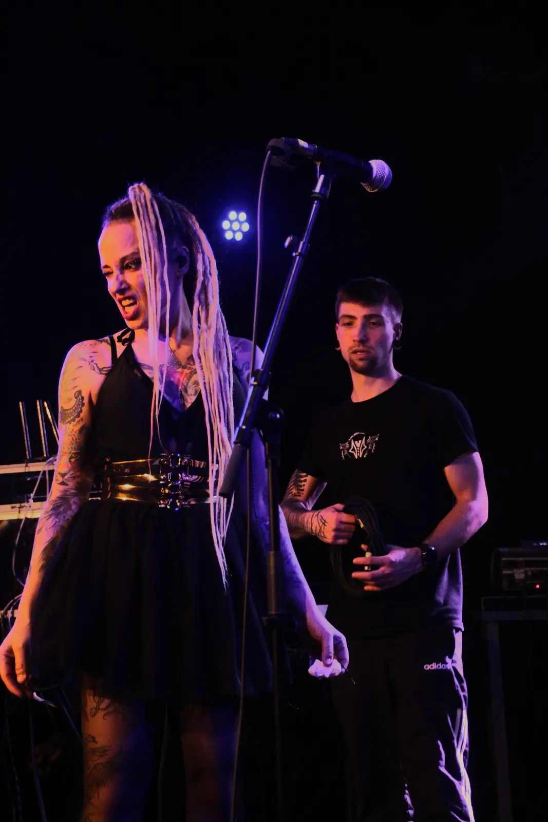 A woman with long white dreadlocks and tattoos, wearing a black dress and gold belt, stands on stage near a microphone…