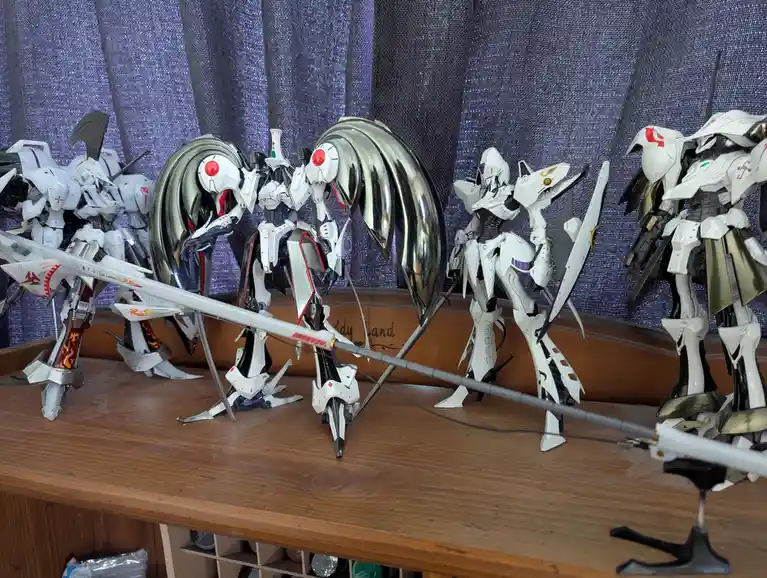 A row of white and silver mecha model kits are displayed on a wooden shelf against a purple curtain. The central mecha has a…