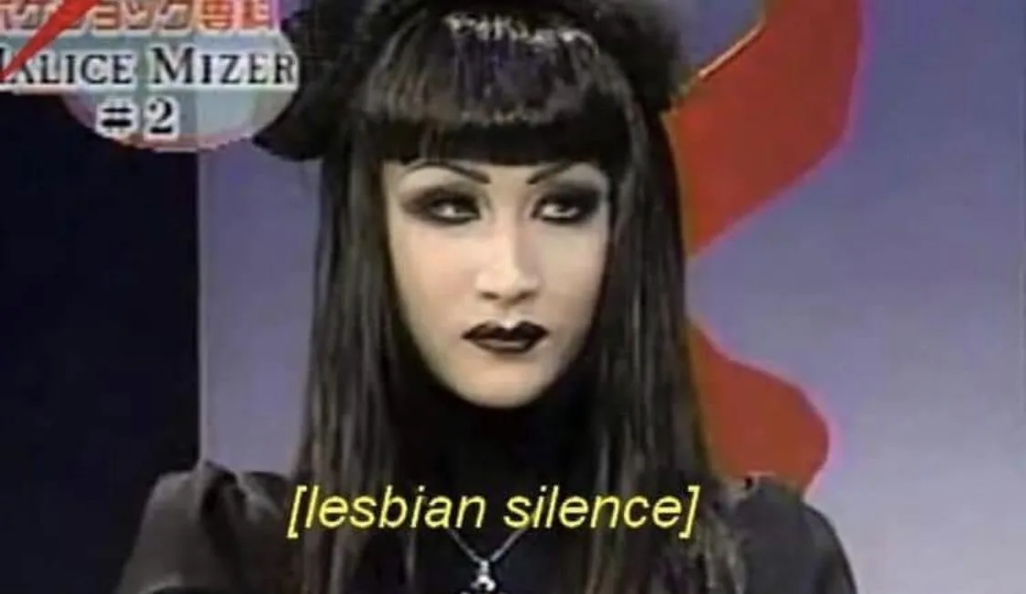 A still image of a woman with dark hair, heavy black eyeliner, and dark lipstick, wearing a black outfit and a black hat…