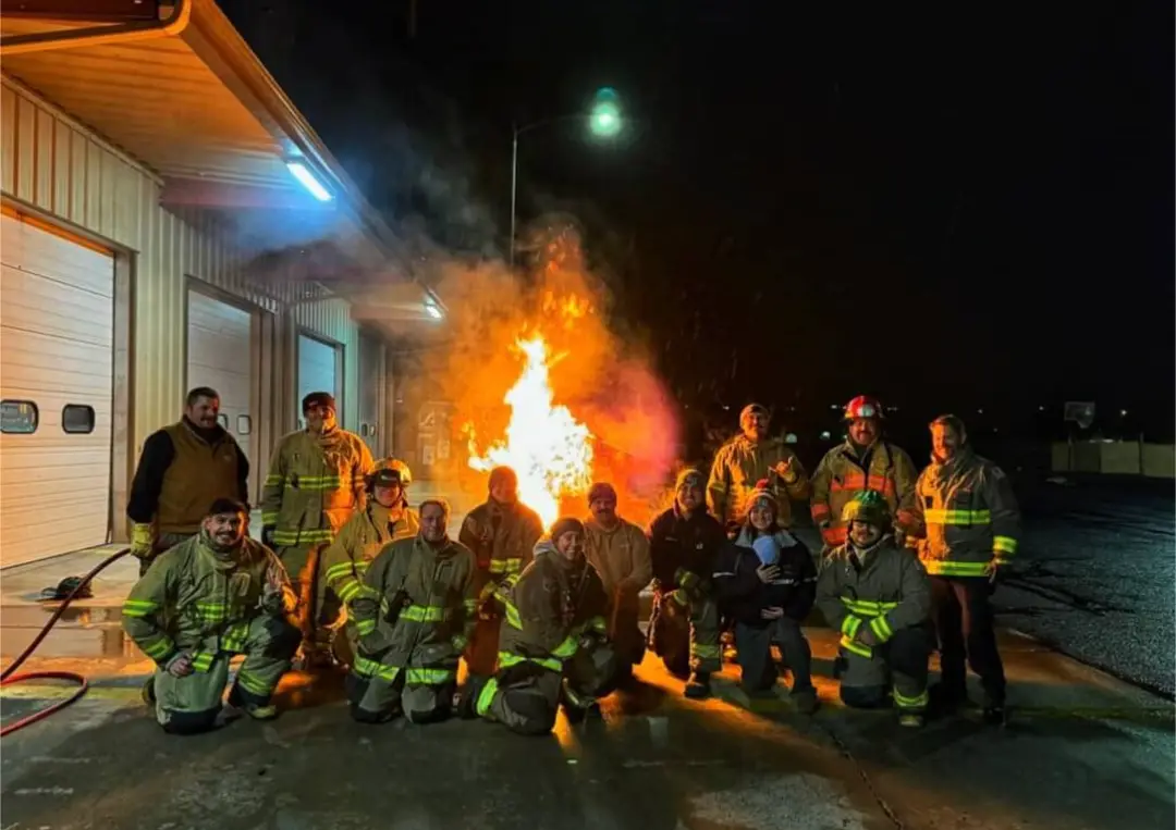 A group of firefighters in full gear pose for a photo at night in front of a large, active fire burning between them and a…