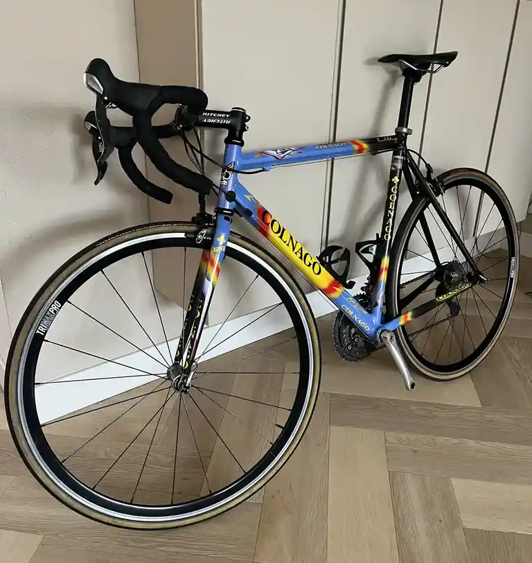 A blue and yellow Colnago road bicycle with rainbow flame decals is leaning against a wall on a wooden floor. The bike has…