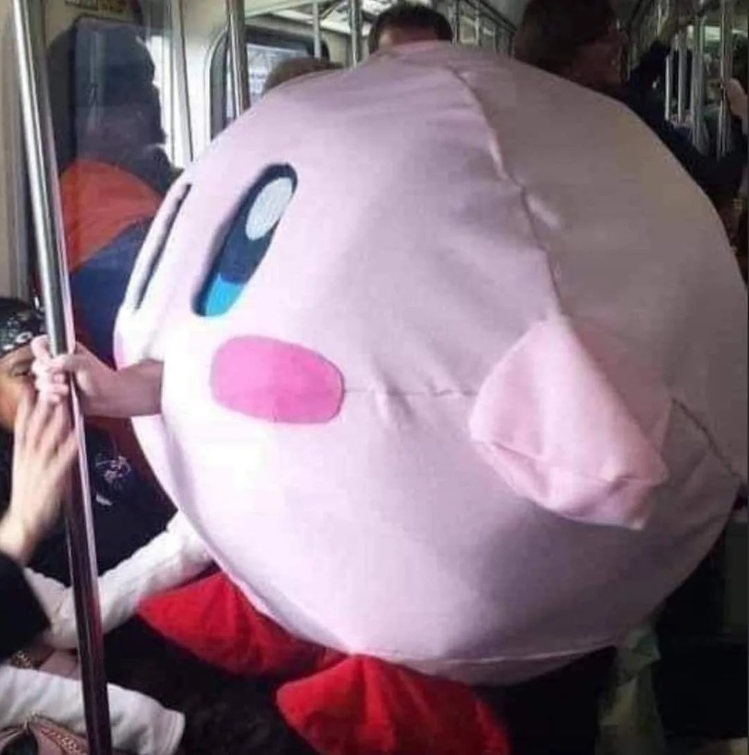 A person wearing a large, pink, round plush costume resembling a cartoon character is standing on a crowded public transit…