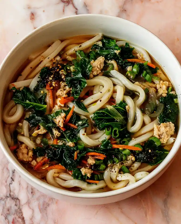 A top-down photo of a white bowl filled with udon noodles, dark broth, chopped kale, shredded carrots, sliced green onions…