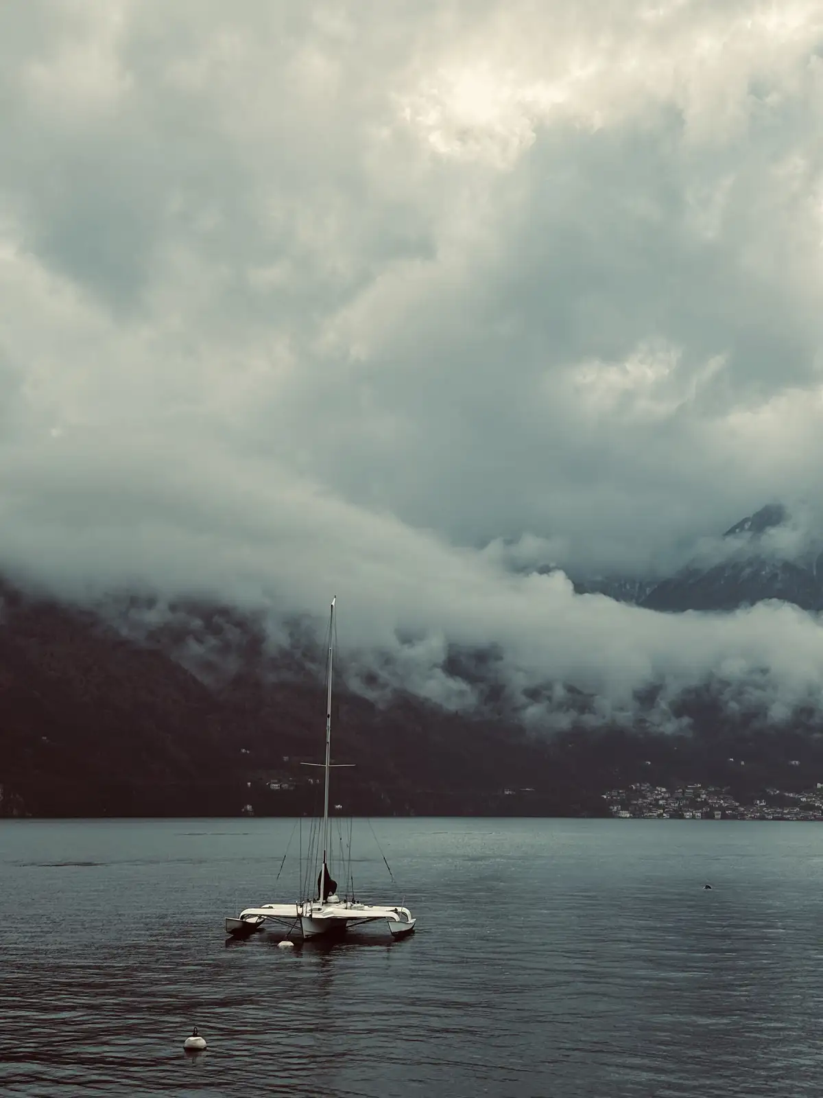 A solitary white catamaran sailboat floats on calm, dark water under a heavy, cloudy sky with misty mountains in the…