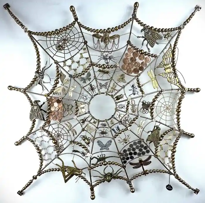 A decorative metal spiderweb art piece features intricate webbing made of brass-colored beads and wires, adorned with…
