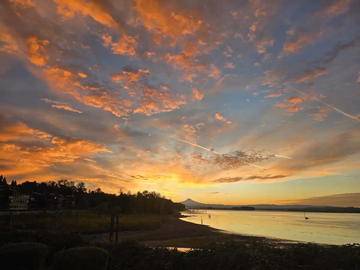 A scenic sunset over a body of water with a distant mountain, featuring vibrant orange and blue clouds, silhouetted trees…