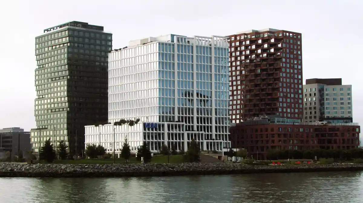 A wide shot of several modern office buildings along a waterfront, with the central white glass skyscraper prominently…