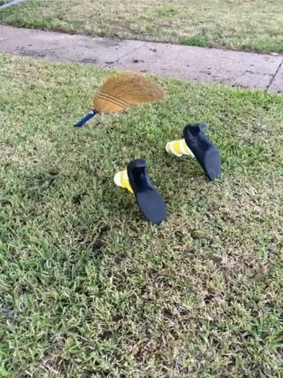 A broom and two black shoes with yellow socks are lying on a grassy lawn, arranged to resemble a face with the broom as hair…