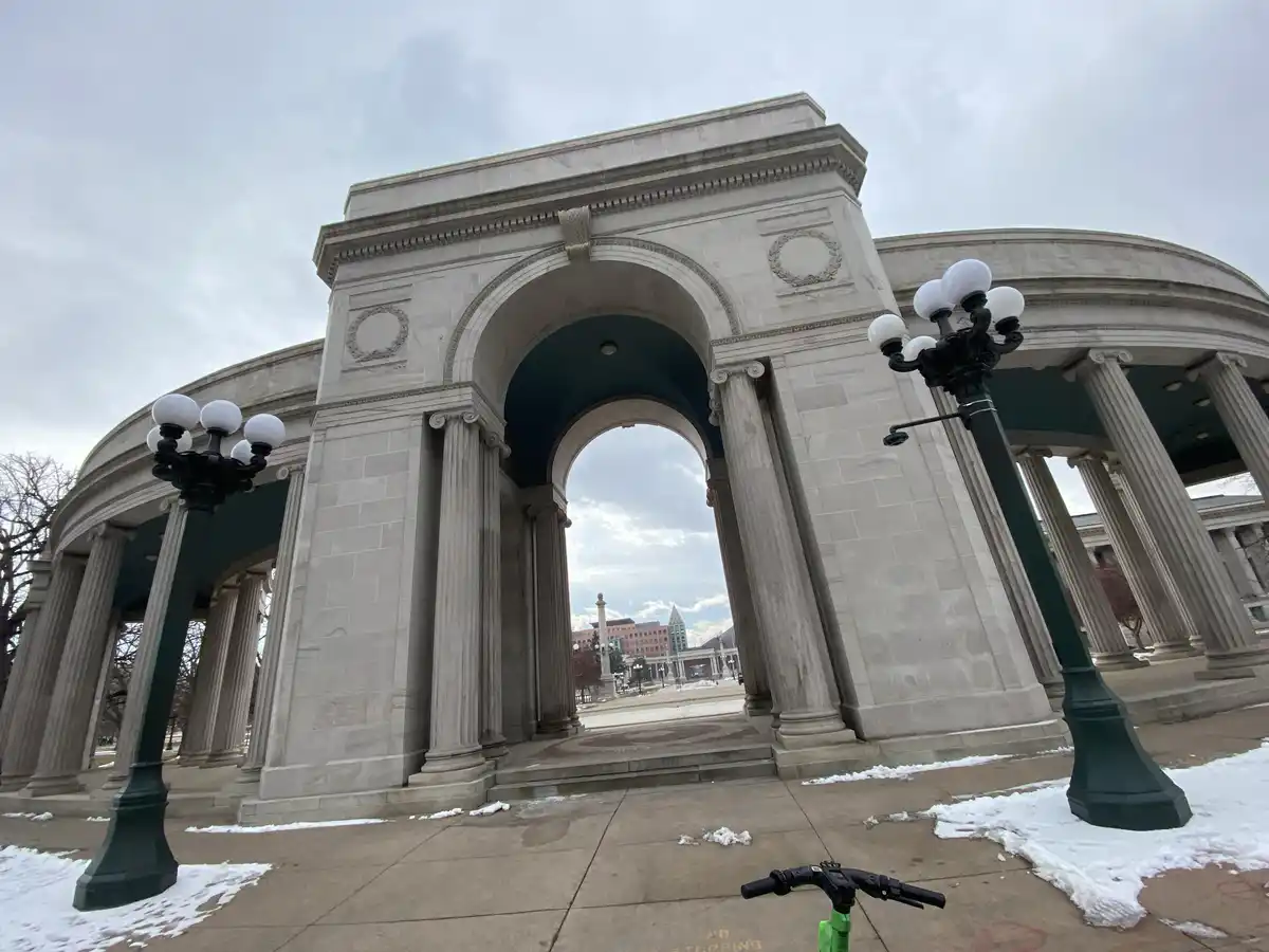 A low-angle photo shows the handlebars of a green electric scooter in the foreground, with patches of snow on the sidewalk…