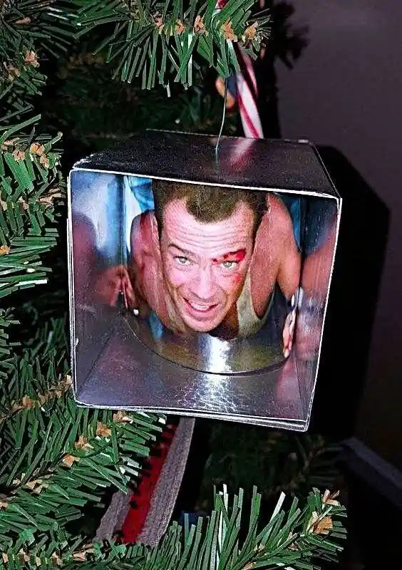 A square metal Christmas ornament hangs on a green pine tree, featuring a printed image of actor Tom Hardy with a bloody eye…