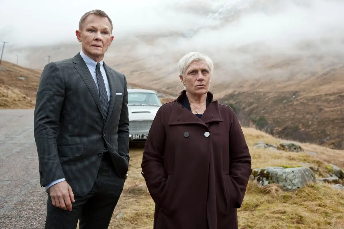 Two people stand on a gravel road in a misty, mountainous landscape with a vintage white car behind them. The man on the…