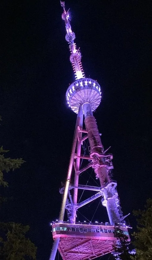A nighttime photo of a tall, illuminated broadcast tower with purple and pink lights, viewed from below against a dark sky…