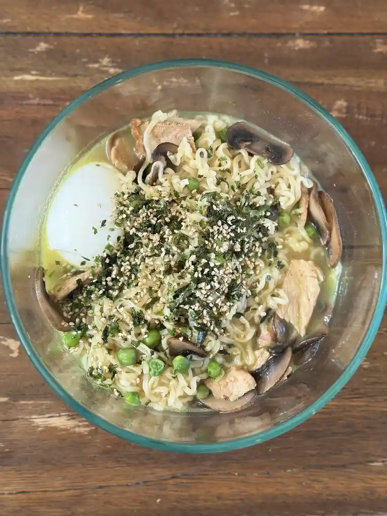 a top-down photo of a clear glass bowl filled with ramen noodles in a light broth, topped with a halved soft-boiled egg…