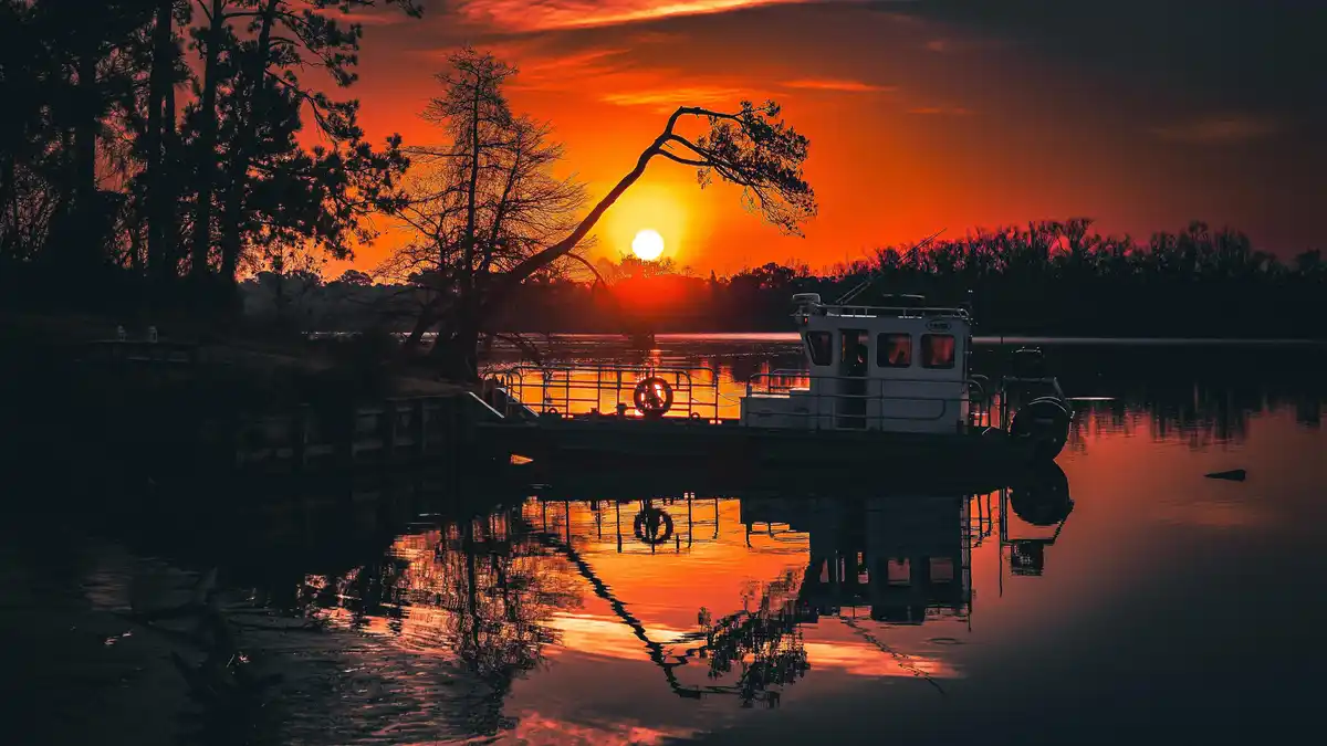 A serene sunset over a calm body of water with a boat docked near the shore. The sky is ablaze with orange and red hues, and…