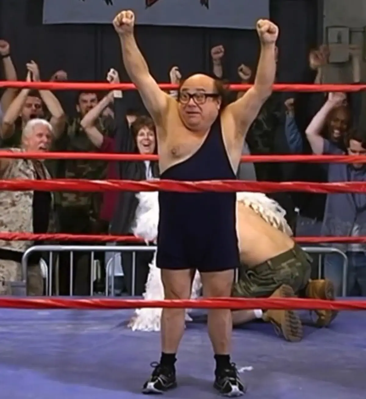 A man in a blue wrestling singlet and glasses stands in a boxing ring with arms raised in victory, while another man in…