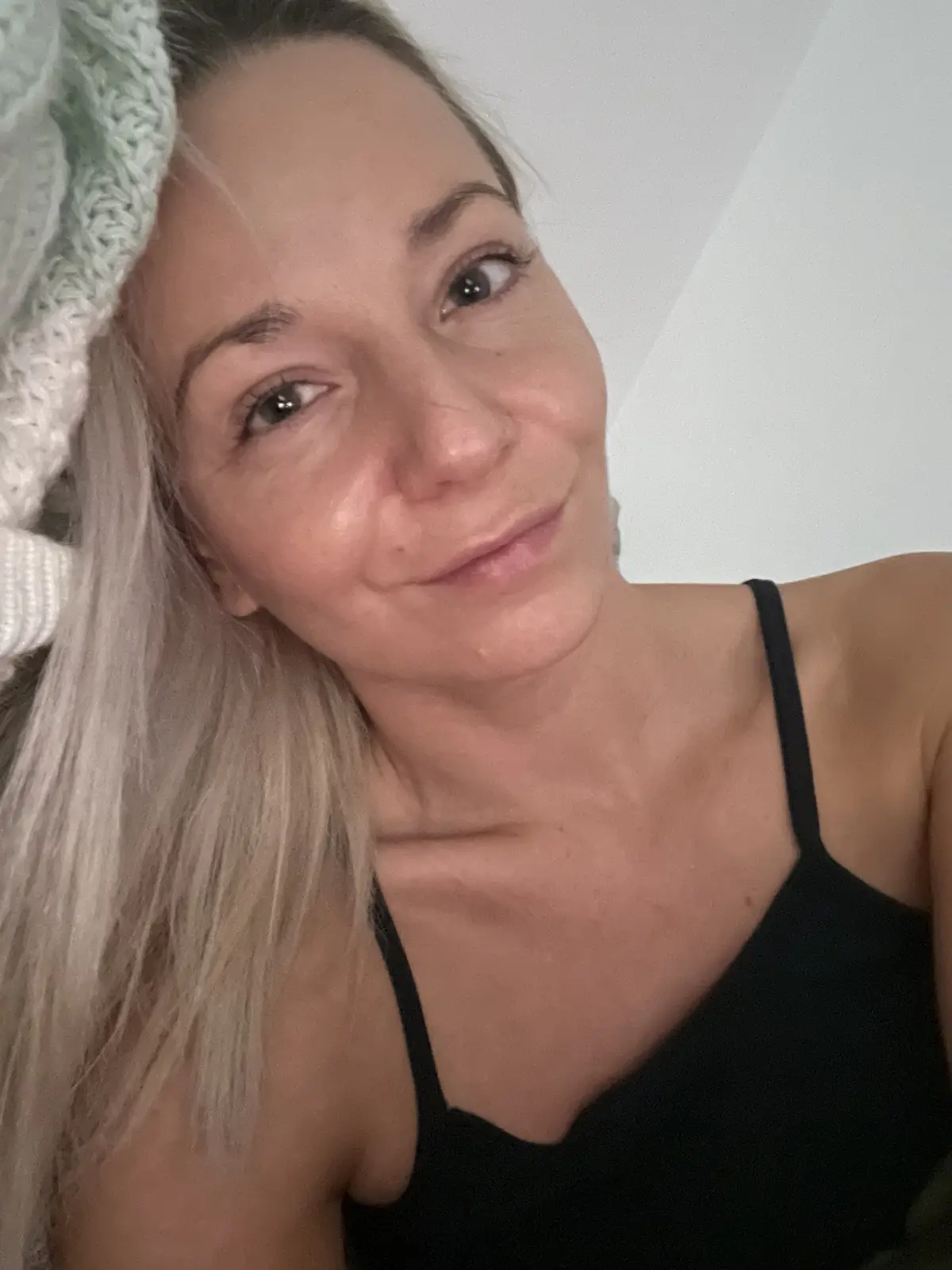 A selfie of a woman with blonde hair wearing a light green knitted head wrap and a black tank top, looking at the camera…