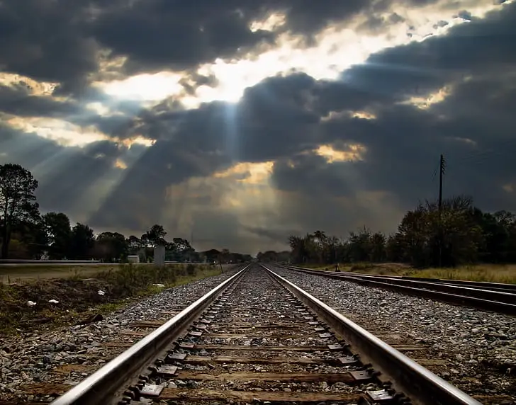 A dramatic photograph of train tracks stretching into the distance under a cloudy sky with sunlight breaking through. The…
