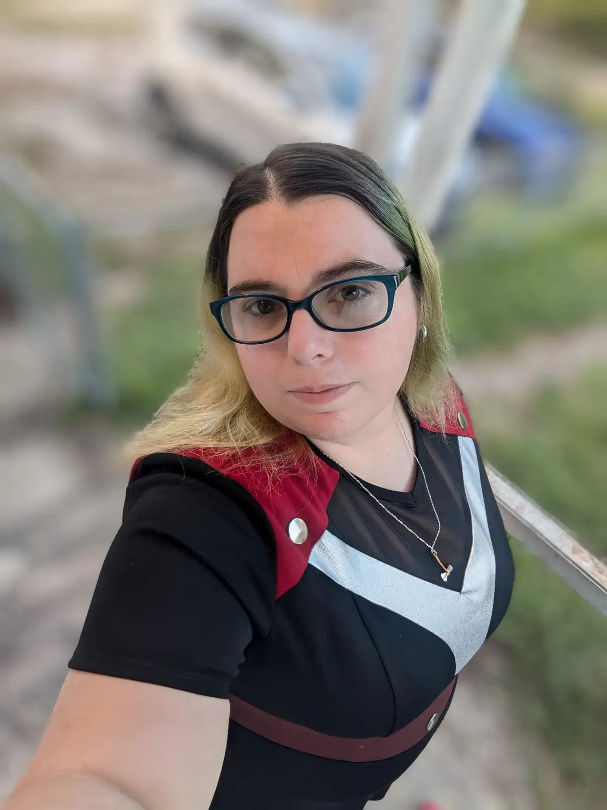 A person with glasses and blonde hair with green highlights takes a selfie wearing a black, red, and silver outfit with…