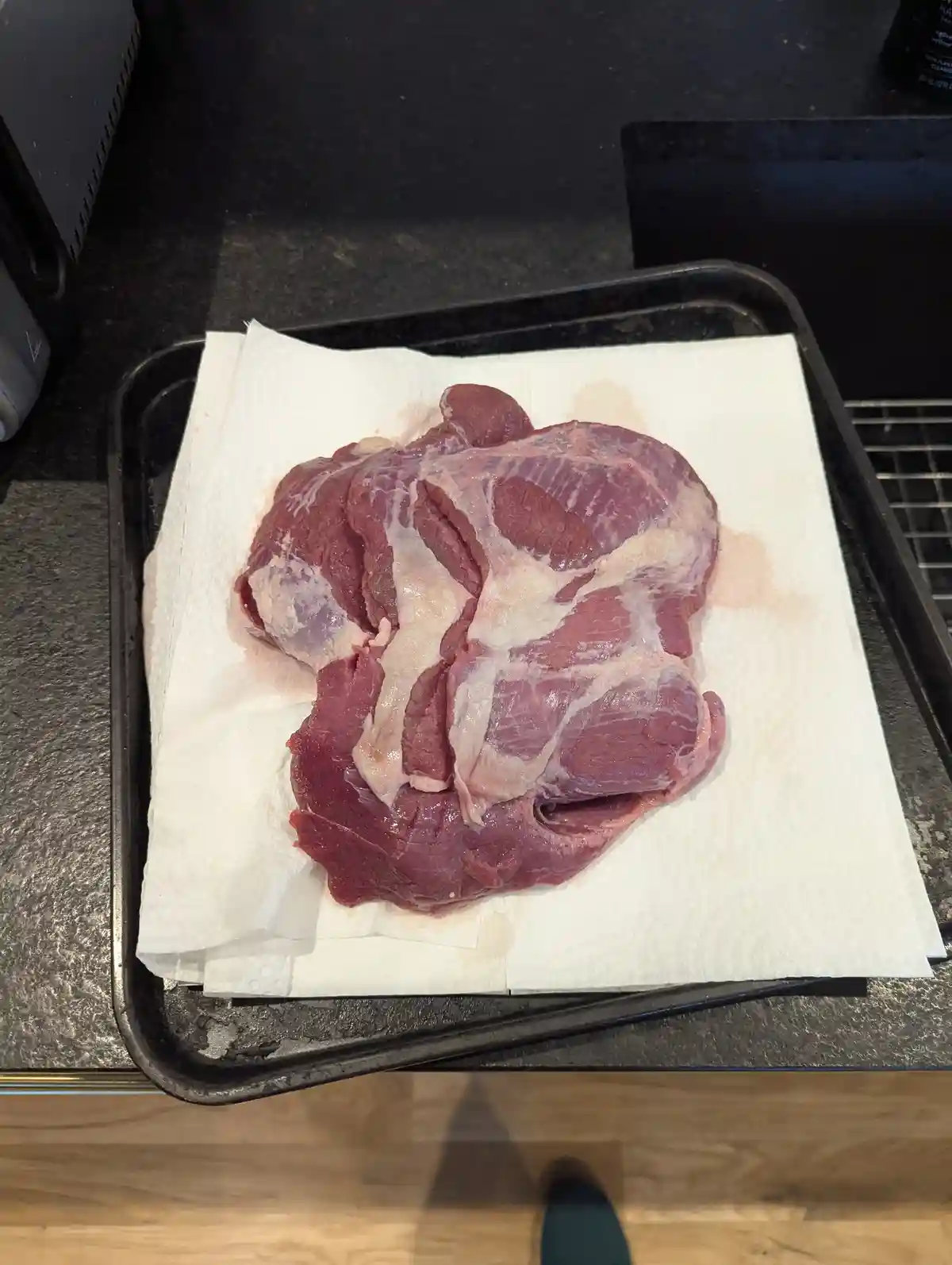 A large, raw piece of red meat with visible fat and connective tissue rests on a white paper towel inside a black baking…