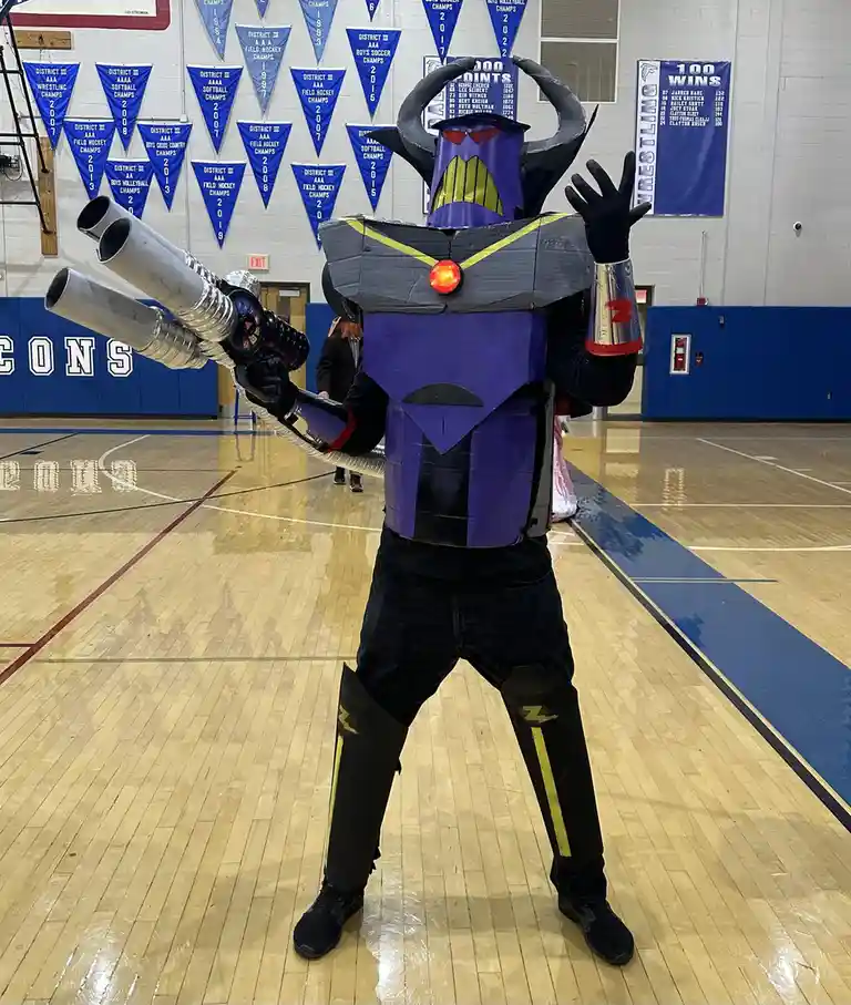 A person in a homemade, cardboard-like Zalgo costume stands in a gymnasium, holding a large, duct-taped weapon. The costume…
