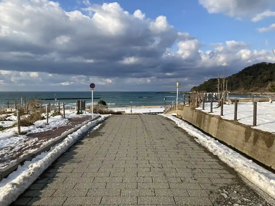 A paved walkway leads toward a beach with snow piled along its edges and on the ground, under a partly cloudy sky with the…