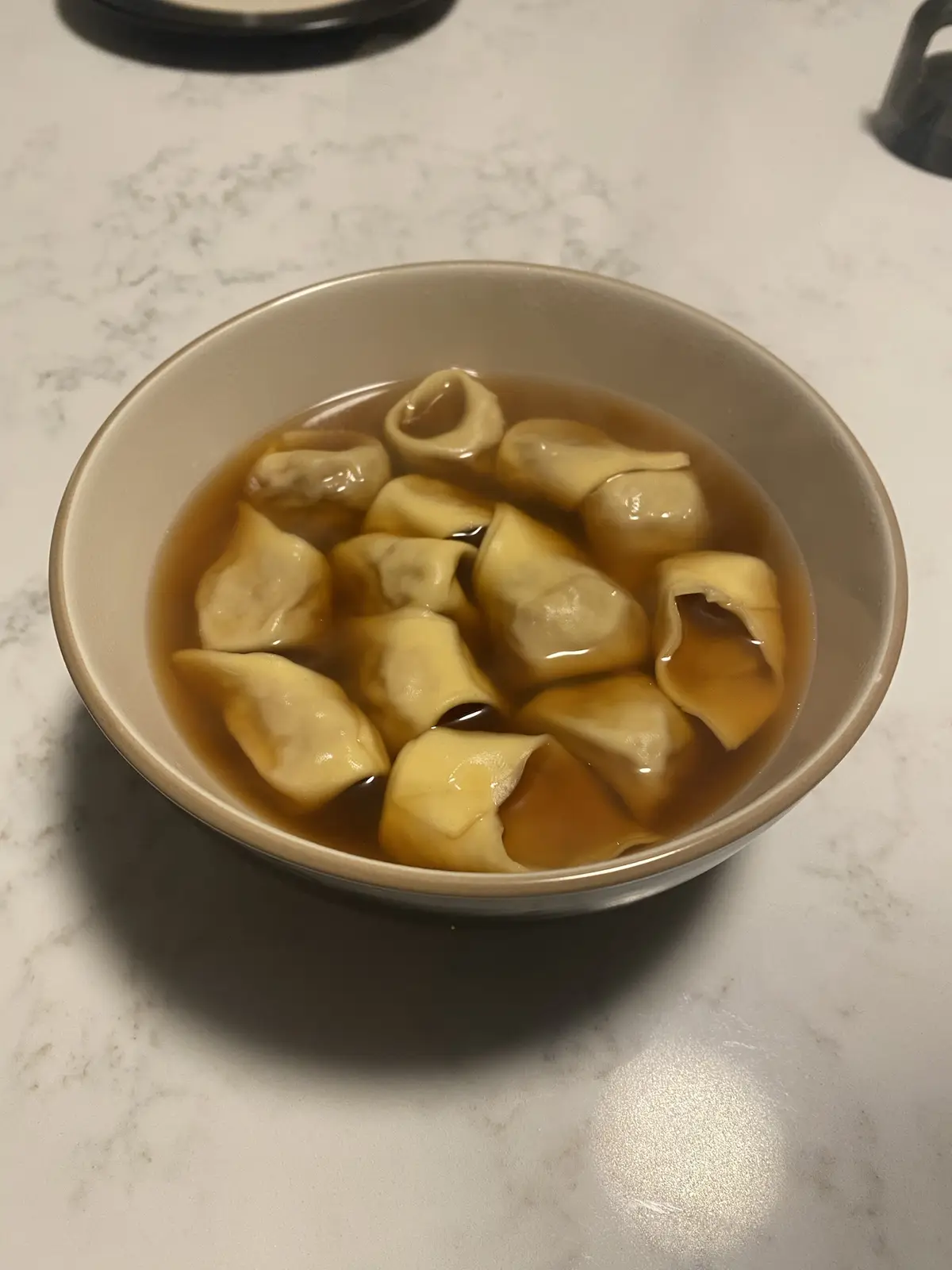 A beige bowl filled with wontons floating in broth sits on a white marble countertop. The wontons are pale yellow and appear…