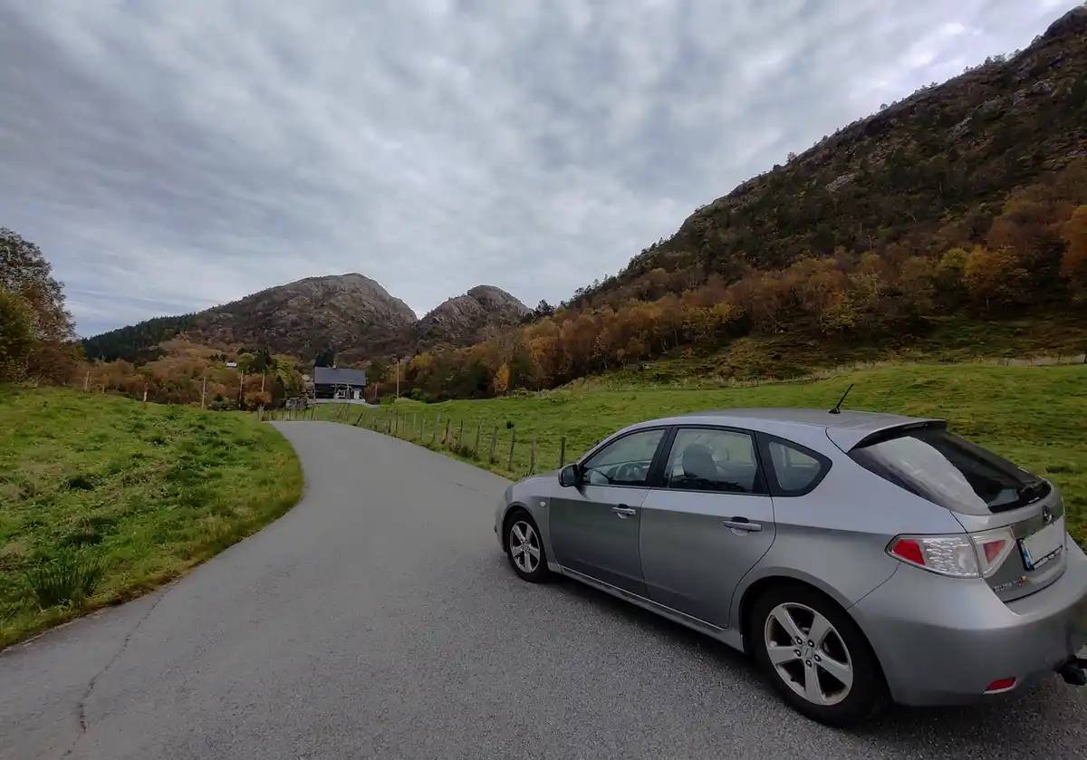 A silver hatchback car is parked on a winding rural road surrounded by green fields and autumn-colored trees with rocky…