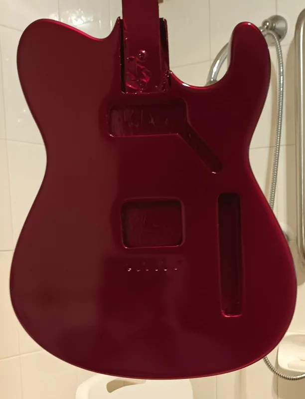 A glossy, deep red electric guitar body is hanging upside down in a bathroom, with a shower head and tiled wall visible in…