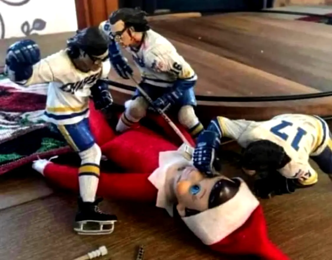 Three miniature hockey player figurines are shown attacking a red-clothed elf doll lying on a wooden floor. The elf has a…