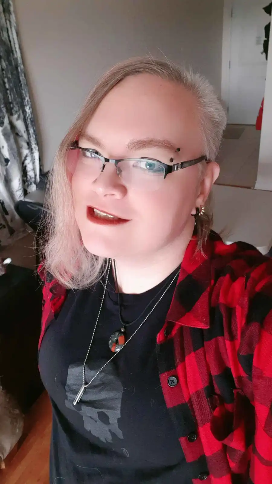 A person with short, light-colored hair and glasses, wearing a red and black plaid shirt over a black t-shirt, smiles…
