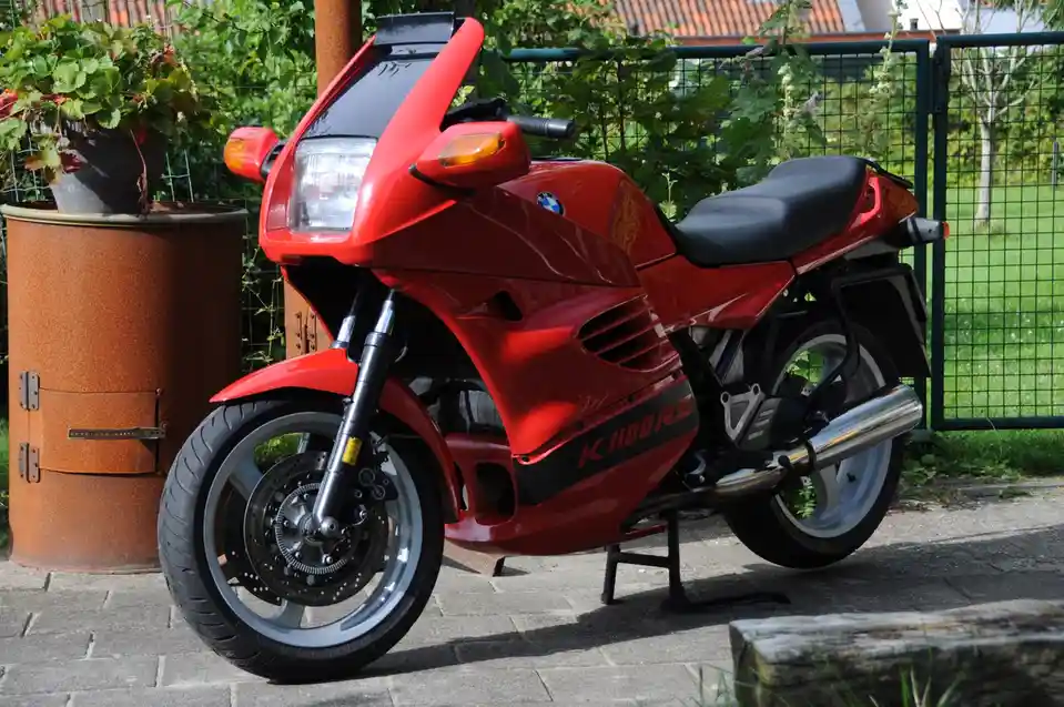 A red BMW K1100RS motorcycle is parked on a paved surface next to a rusty metal barrel with a potted plant on top, with…