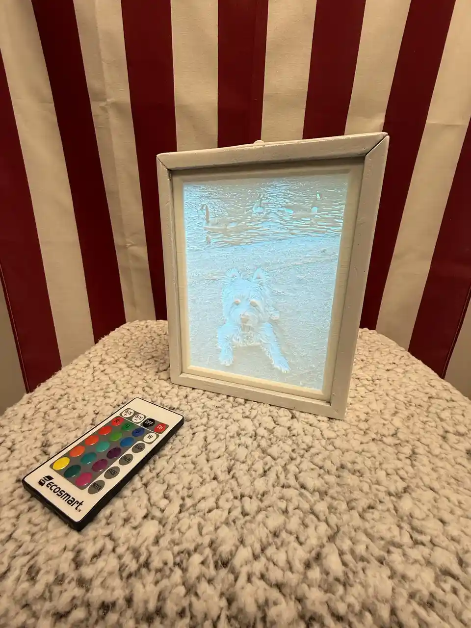 A white picture frame sits on a fluffy beige surface, projecting a glowing image of a small dog in front of water with…