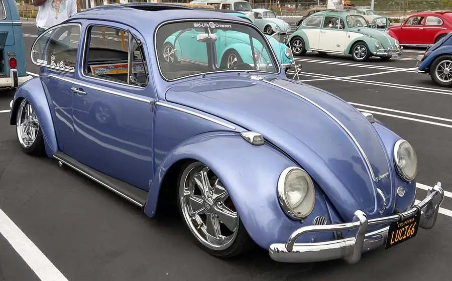A blue Volkswagen Beetle with chrome rims and a chrome bumper is parked in a lot with other Beetles. The windshield has a…