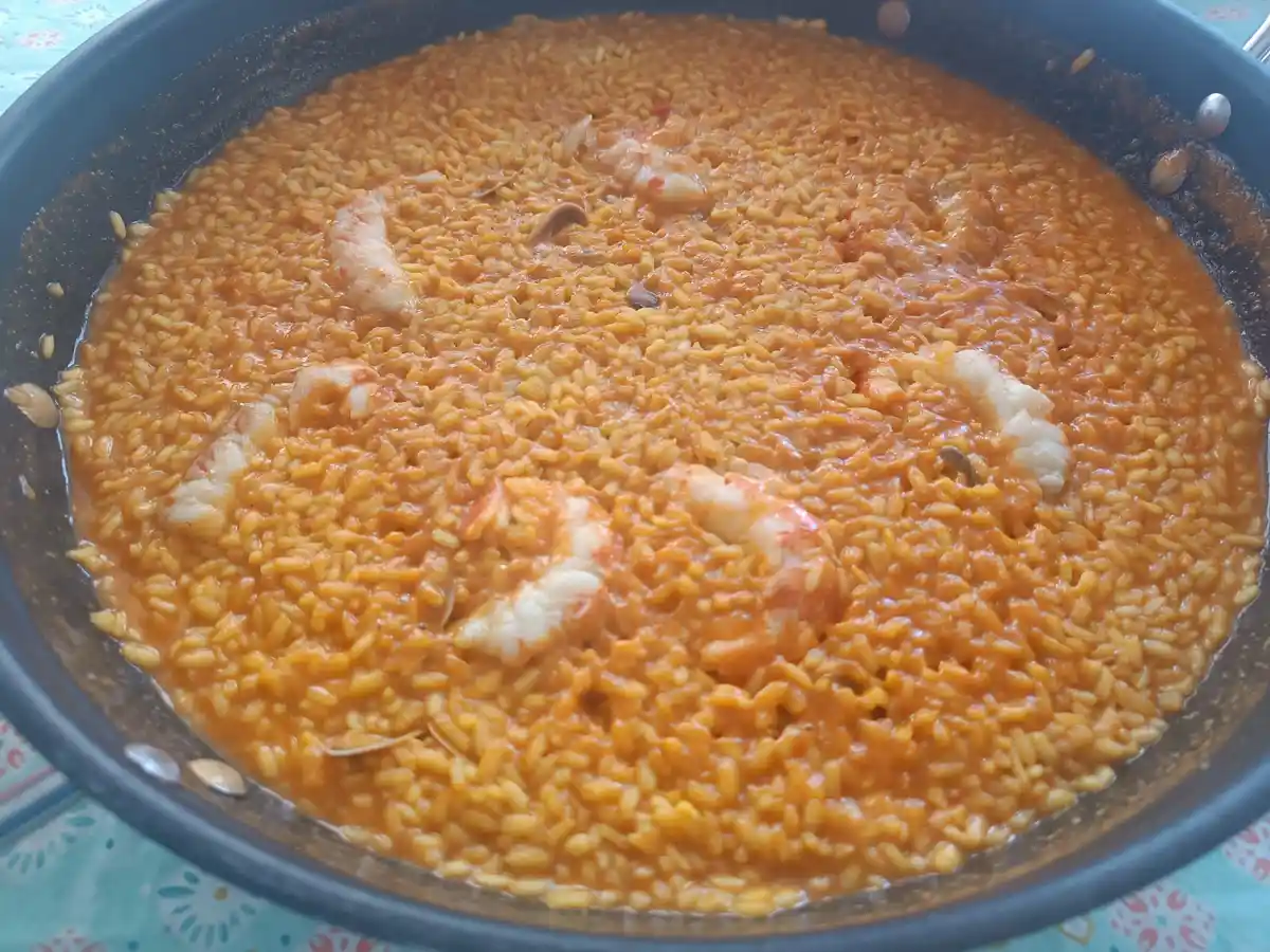 A close-up photo shows a dark pan filled with orange-colored rice dish, likely paella, containing visible shrimp and clams…