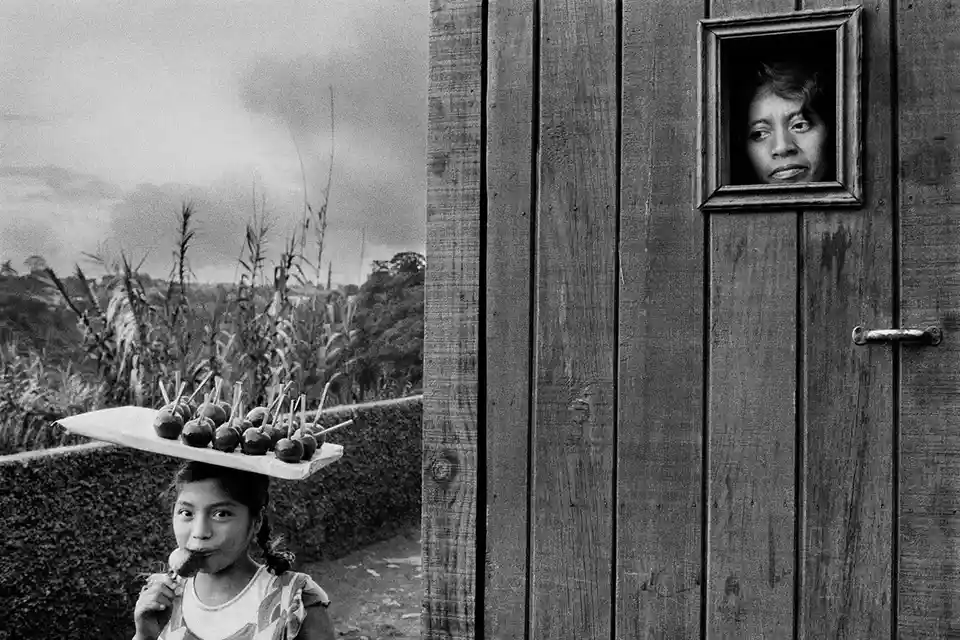 A black-and-white photo shows a young girl balancing a tray of lollipop-like treats on her head while eating one, with a…