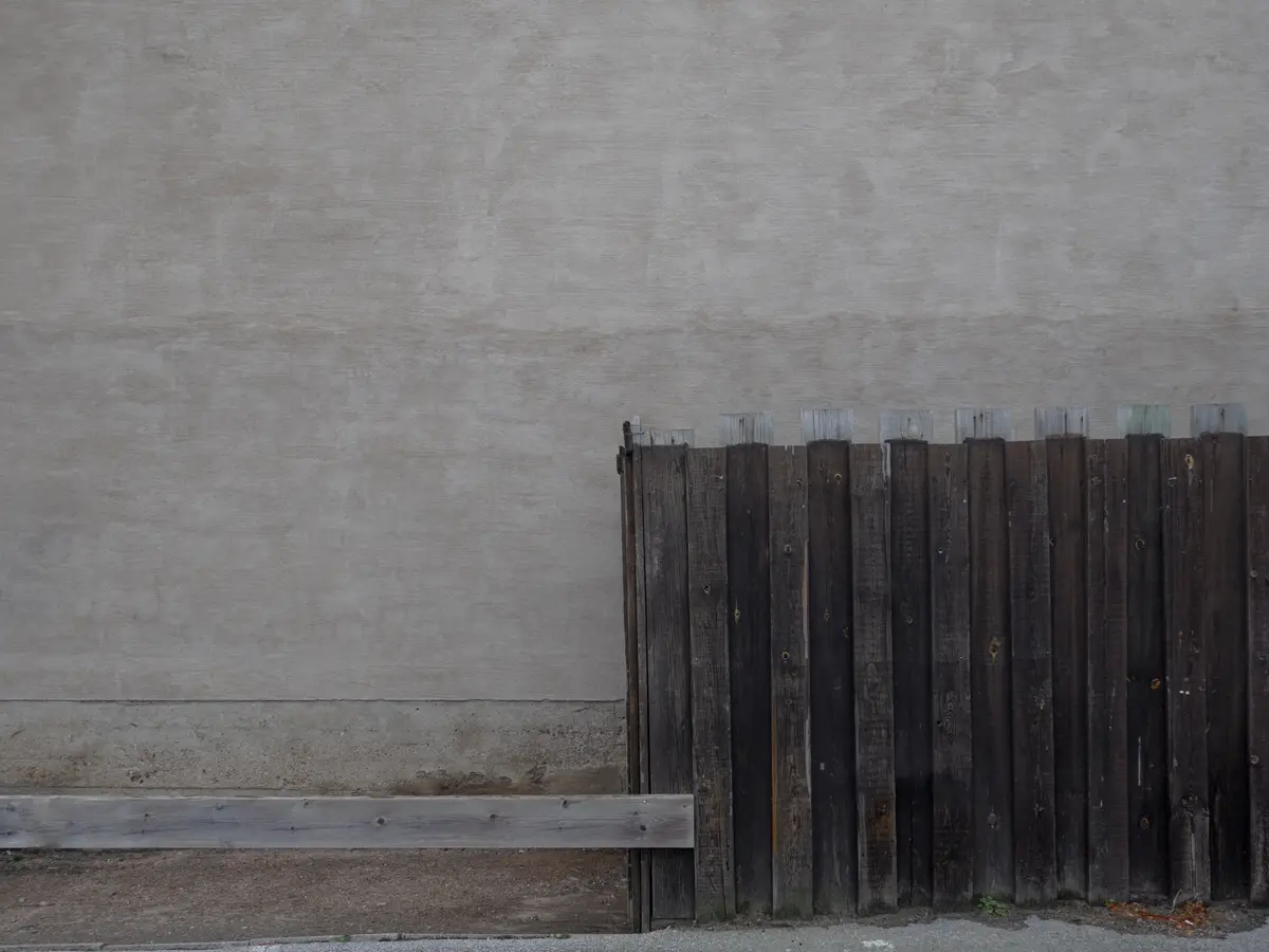 A weathered wooden fence stands against a plain concrete wall, with a horizontal wooden plank at its base. The image is…