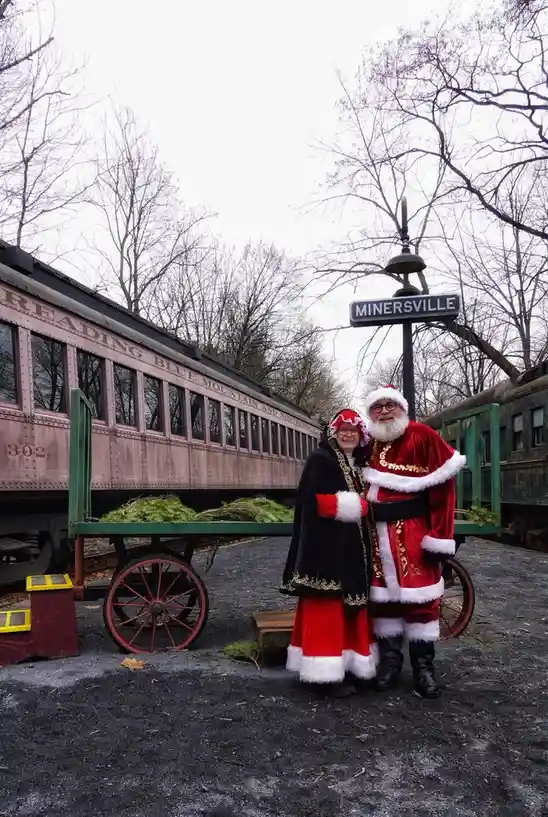 Two people dressed as Santa and Mrs. Claus pose for a photo in front of a vintage train car labeled 'READING BLUE MOUNTAIN…