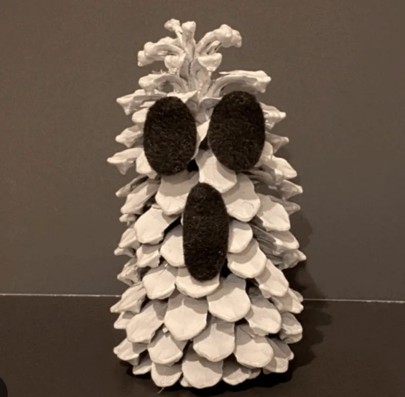 A white-painted pinecone has three black fuzzy oval shapes glued to it to resemble a ghost's eyes and mouth. The pinecone…